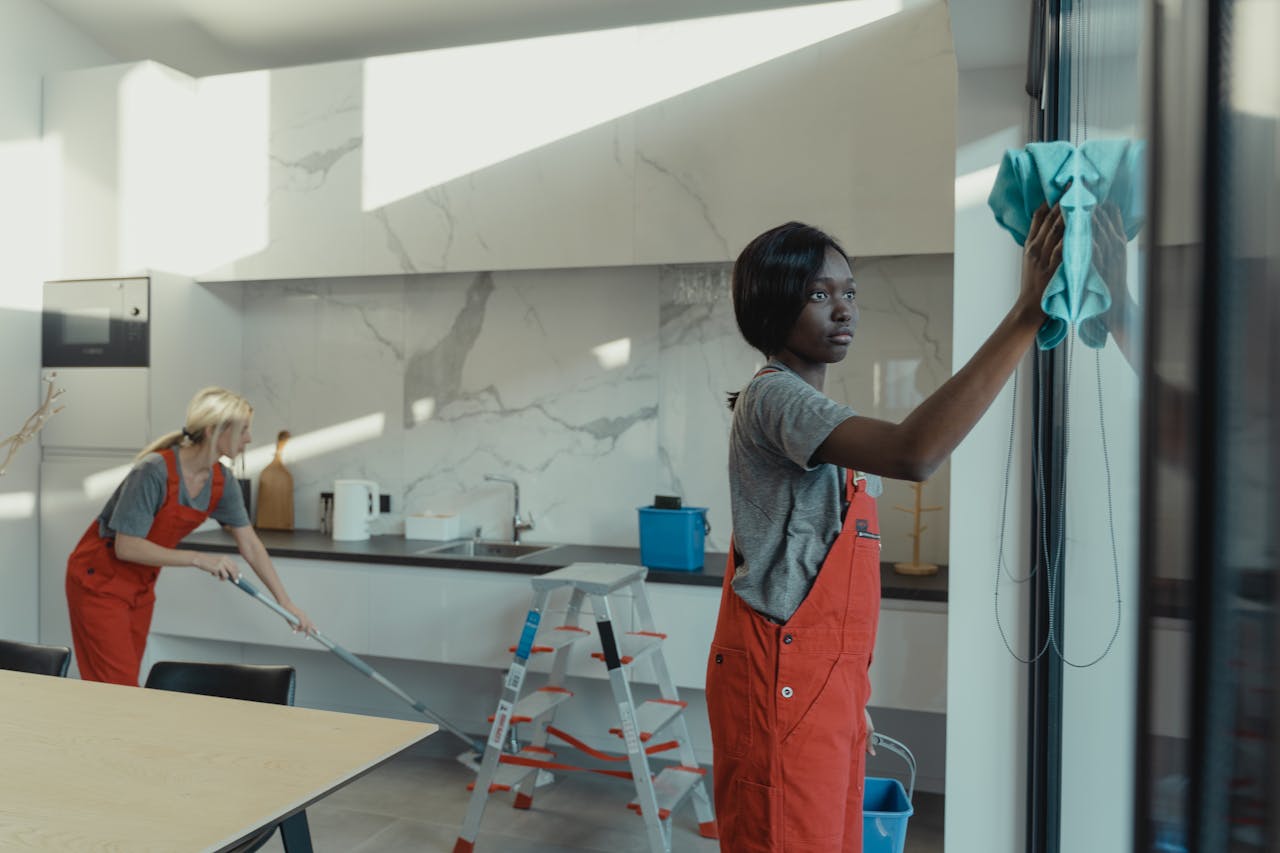 creative-03 Two women cleaning a modern kitchen, wiping glass surfaces and mopping the floor.
